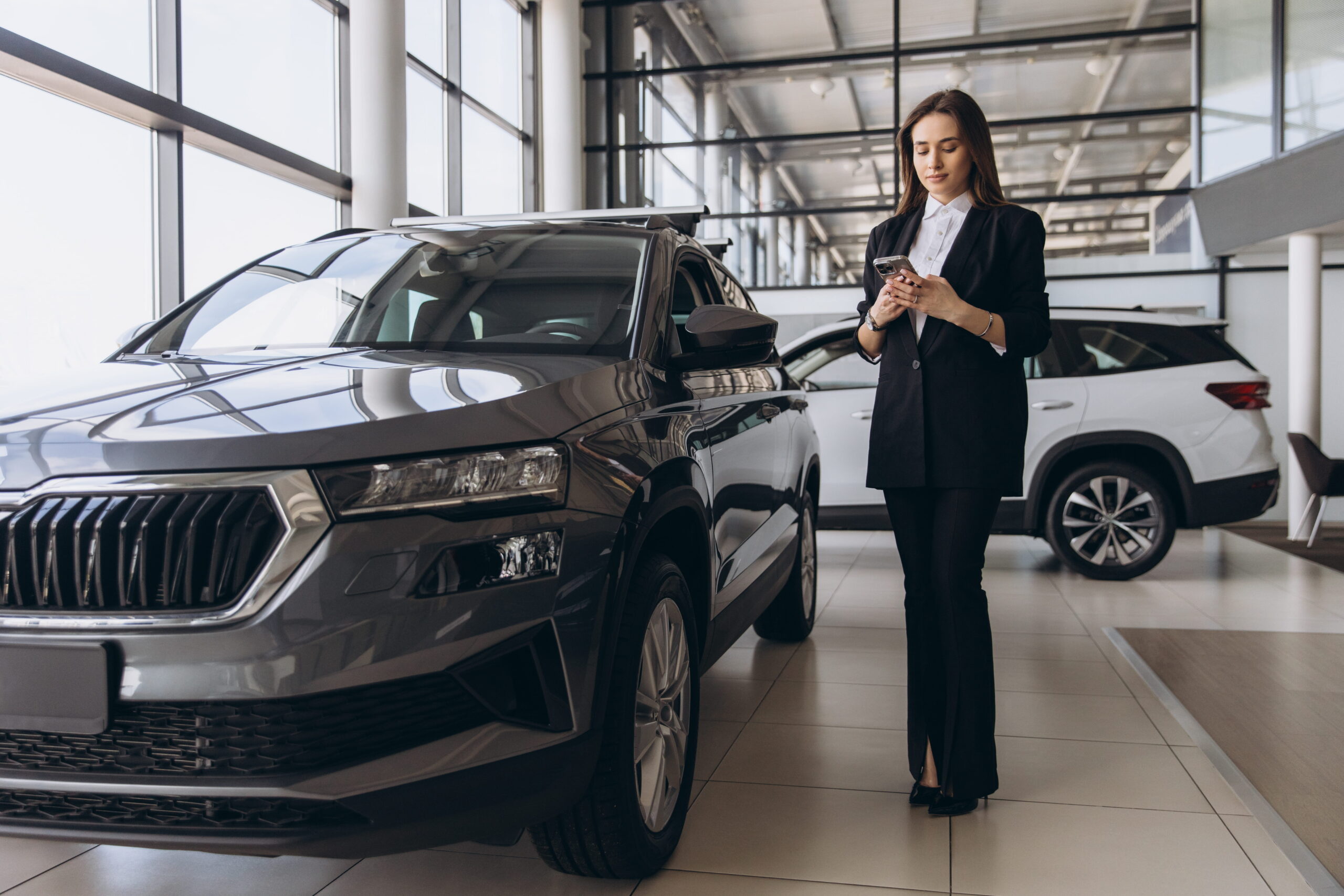 Female automotive manager at car dealership reviewing car specs on phone.